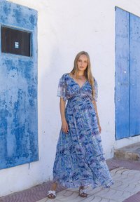 Robe maxi en tissu à imprimé floral bleu avec des manches courtes flottantes, un décolleté en V et une taille cintrée. Présentée devant un mur blanc et des portes bleues.