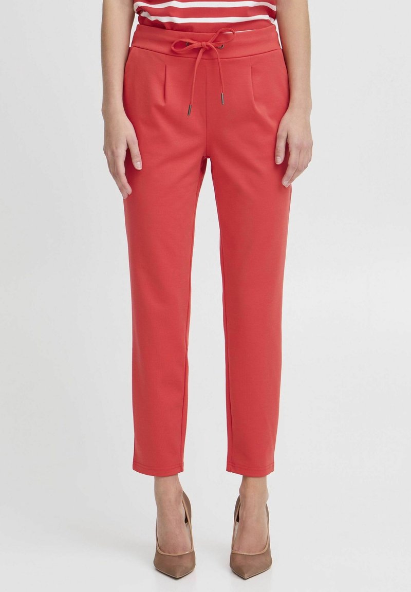 Femme portant un pantalon taille haute corail avec un cordon de serrage et des talons hauts marron à bout pointu, debout devant un fond blanc uni.