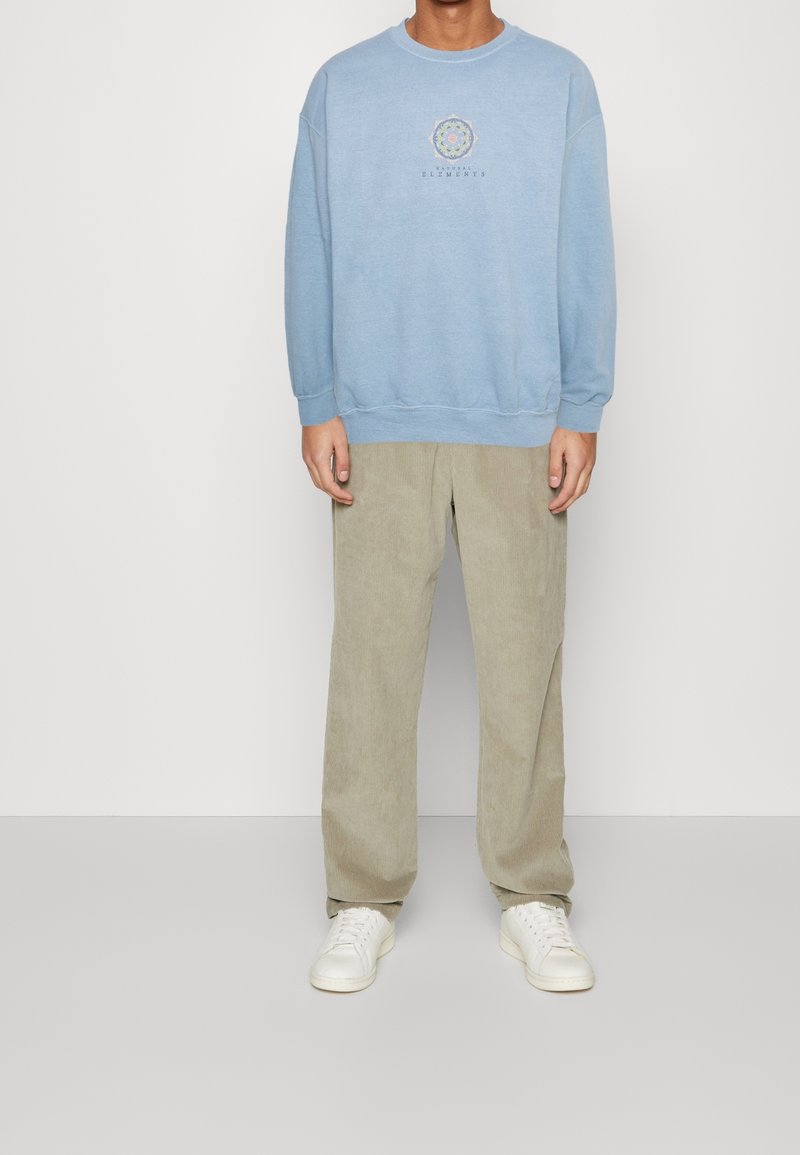 Ljusblå sweatshirt med rund halsringning och subtil grafisk design på bröstet. Matchad med beige kordflorsbyxor och vita sneakers.