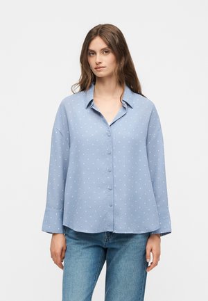 Νεαρή γυναίκα που φορά ανοικτό μπλε πουκάμισο με μακριά μανίκια και πουά σχέδιο και μπλε τζιν μπροστά από απλό φόντο.