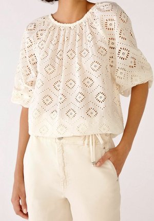 Kobieta w białej bluzce z ażurowej koronki z bufkami, schowanej w spodniach z wysokim stanem w kolorze kremowym, z ręką w kieszeni.