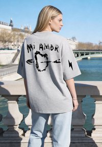Jeune femme portant un T-shirt gris oversize avec « Mi Amor » et un motif de cœur percé, se tenant près d’une rambarde en pierre au bord d’une rivière en ville.