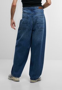 Pantaloni in denim a gamba larga blu scuro, con due tasche posteriori, una vita con passanti per cintura e una toppa in pelle sul retro.