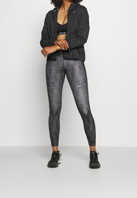 Tenue de sport comprenant une veste noire, des leggings noirs à motifs avec un texte blanc, et des baskets noires. Design ajusté et matériau léger.
