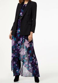 Musta bleiseri, jossa on kaksoispainikkeet, violetin ja vihreän kukkamekon päällä. Mekossa on kerroksellinen, läpikuultava, röyhelöhelma ja nilkkapituiset mustat saappaat.