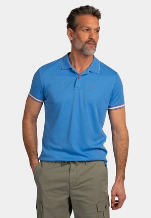 Man met grijze baard die een blauw poloshirt met korte mouwen en kaki cargobroek draagt, kijkt opzij met zijn handen gedeeltelijk in zijn zakken.