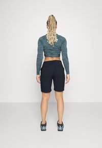 Haut à manches longues en teal et short noir, avec une coupe ajustée et une texture lisse, poches à l'arrière et chaussures de sport.