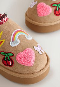 Tan suede slippers with embroidered patches: a rainbow, pink heart, cherries, and butterflies. Textured accents and a round toe design.