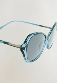 Okuliare s veľkými, zaoblenými šošovkami v aqua farbe; elegantné priesvitné rámy; strieborné detaily na pántoch.