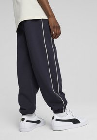 Pantaloni della tuta blu navy con strisce laterali bianche, polsini a costine e vestibilità rilassata, abbinati a sneaker bianche classiche con dettagli neri.