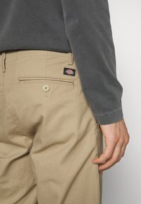 Pantalon beige Dickies avec poche boutonnée au dos, porté avec une chemise à manches longues gris foncé, main détendue sur le côté.