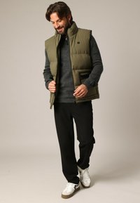 Olivengroene mouwloze gewatteerde bodywarmer over een donkergrijze trui, zwarte broek en witte sneakers met grijze accenten. Eenvoudig ontwerp, zichtbare zak.