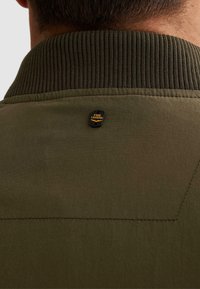 Zadní část krku osoby nosící olivově zelenou bundu s žebrovaným límcem a malou černou značkou s logem PME Legend.