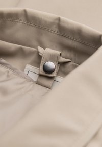 Tessuto beige con una texture liscia presenta una chiusura a scatto e un dettaglio ad anello, mettendo in mostra cuciture pulite e una leggera lucentezza.