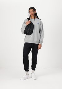 Sweatshirt cinza com logótipo azul, mala tiracolo preta, calças de fato pretas e ténis brancos. Tecido suave, corte relaxado e aspeto casual.