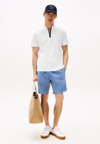 Jonge man in een witte polo met ritssluiting, blauwe shorts, witte sneakers, een marineblauwe pet, en houdt een tan tote bag vast met witte handvatten.