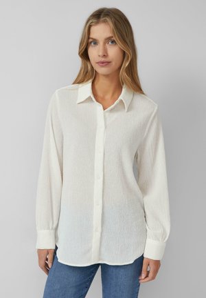 Junge Frau mit blonden Haaren, die eine strukturierte weiße Bluse mit langen Ärmeln und blauen Jeans trägt, steht vor einem schlichten Hintergrund.