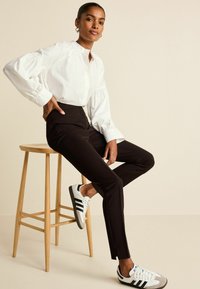 Witte blouse met lange mouwen en gathered manchetten, gecombineerd met donkerbruine-tailored broek en witte sneakers met zwarte strepen en grijze accenten.