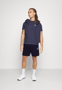 Ensemble de sport bleu marine composé d'un t-shirt à manches courtes et d'un short assorti. Le t-shirt présente un logo, et le mannequin porte des baskets blanches.