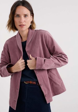 Woman adjusting a mauve buttoned jacket over a black top, wearing a braided brown belt and navy skirt against a plain light background.