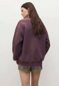 Sudadera de color púrpura oscuro, de corte relajado, con puños de canalé. Combinada con pantalones cortos de satén con borde de encaje verde. Vista trasera del conjunto.