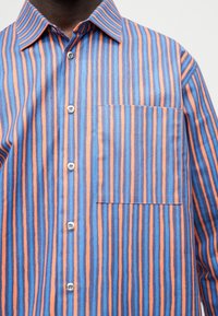 Man draagt een overhemd met verticale strepen in blauw, oranje en marineblauw, met een borstzak aan de linkerkant.