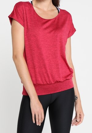 Kvinde iført en loose-fit rød kortærmet top og sorte leggings, der viser en tatovering på hendes venstre håndled mod en ensfarvet baggrund.