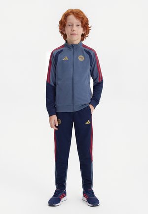 Copil cu păr roșu stă îmbrăcat într-un trening sportiv albastru și bleumarin cu dungi roșii și pantofi sport bleumarin asortați cu accente roșii.