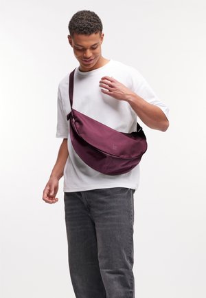 Bordeauxfarbenes Umhängetasche aus Stoff mit einem geschwungenen Design. Verfügt über einen einzelnen Träger und ein kleines Logo. Wird quer über den Körper getragen.