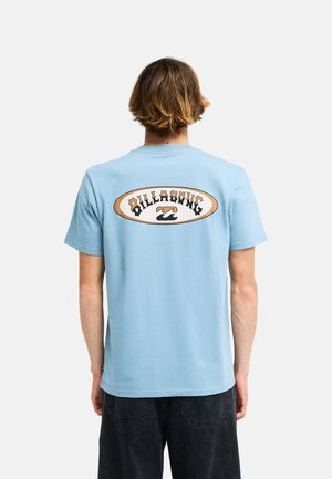 Uomo con capelli corti castani che indossa una maglietta Billabong azzurra chiara e pantaloni neri, visto da dietro su uno sfondo semplice.