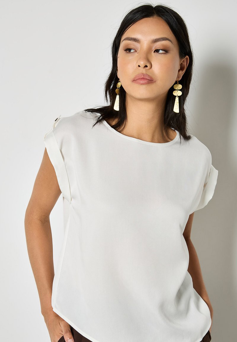 Femme portant un chemisier blanc ample aux manches retroussées, avec de longues boucles d'oreilles pendantes en or, regardant vers sa gauche sur un fond uni.