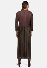 Brązowa zamszowa krótka kurtka z długimi rękawami, noszona z plisowaną długą ciemnozieloną spódnicą, dopełniona brązowymi butami za kolano.