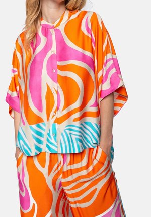 Vrouw draagt een kleurrijk los knoopoverhemd en bijpassende shorts met feloranje, roze, blauwe en witte abstracte patronen.