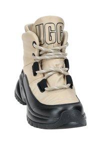 Bota de senderismo acolchada en beige con suela de goma negra, cordones plateados y un prominente logo de UGG. Presenta un diseño de soporte alto para el tobillo.
