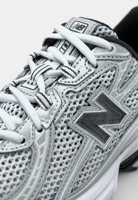 Grauer und weißer Sportschuh mit einem Obermaterial aus Mesh, Kunststoff- und Gummielementen, mit einem auffälligen schwarzen "N"-Logo und Schnürsenkeln.