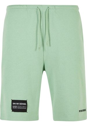 Mäta zelené bavlnené kraťasy s elastickým pásom, šnúrkou a čiernou nášivkou na ľavom stehne s bielym textom. Hladká textúra.