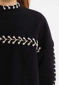 Mustat neuleet, jossa on korkea kaulus ja kermanvärisiä nauhakaarroksia rinnassa ja hihoissa. Pehmeä rakenne, ylipitkä malli, rento muotoilu.