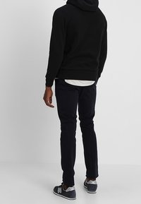 Muž stojící zády, oblečený v černé mikině s kapucí, bílé košili, tmavých kalhotách a šedých teniskách New Balance na jednolitém pozadí.