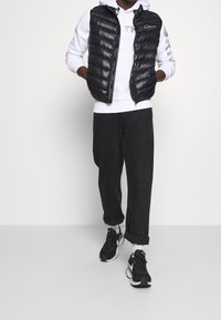 Schwarze gesteppte Weste mit glänzendem Finish, getragen über einem weißen Hoodie, kombiniert mit schwarzen, locker sitzenden Hosen und schwarzen Sneakern.