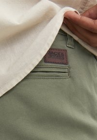 Main levant une chemise beige pour révéler un pantalon olive avec un patch de marque marron JACK & JONES près de la poche arrière.