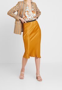 Kvinde iført en sennepsfarvet satin midi-nederdel, blomsterbluse, stribet blazer draperet over skuldrene og hvide hæl-sandaler, stående med hænderne på hofterne.