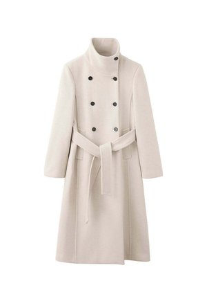 Femme aux longs cheveux bouclés portant un manteau beige à double boutonnage avec un col montant et une ceinture, face à un fond uni.