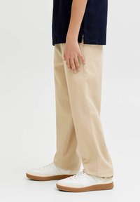 Personne portant un pantalon beige et des baskets blanches avec des semelles en gomme, se tenant devant un fond clair uni.