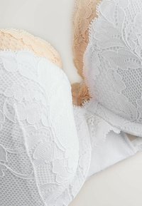 Biustonosz z białej koronkowej tkaniny i cielistego materiału, zdobiony kwiatowymi wzorami, z fiszbinami, gładką teksturą i delikatnymi wykończeniami.