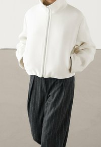 White zip-up jacket with a high collar and long sleeves, paired with dark pinstriped trousers. Soft, textured fabric with a relaxed fit.
