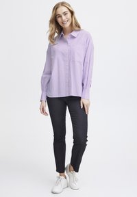 Light purple striped button-up shirt with a chest pocket, paired with dark fitted pants and white sneakers. Smooth fabric, relaxed fit.