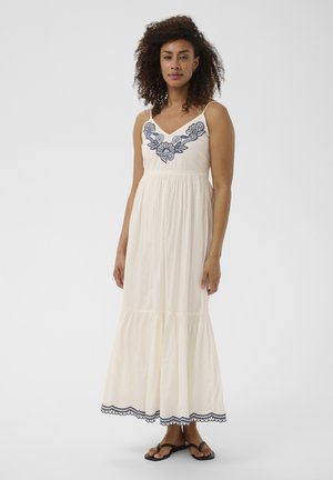 Femme aux cheveux bouclés portant une robe maxi blanche sans manches avec une broderie florale bleue et des sandales noires, debout sur fond blanc.