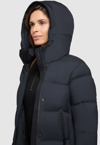 Donkerblauwe pufferjack met een capuchon, voorzien van een ritssluiting aan de voorkant, gestructureerde stof en zwarte accenten. Inclusief zijzakken en een getailleerd ontwerp.