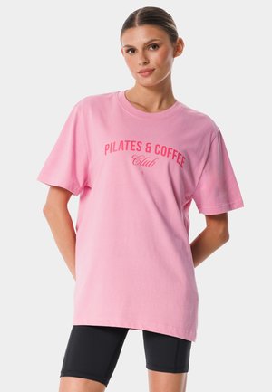 Kobieta z zaczesanymi do tyłu włosami, ubrana w luźny różowy t-shirt z napisem „Pilates & Coffee Club” oraz czarne spodenki kolarskie, stojąca z rękami za plecami.