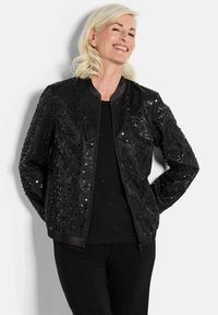 Veste bomber noire avec sequins, dotée d'une fermeture éclair, d'un col rond et de poignets côtelés. Portée sur une chemise noire texturée et un pantalon.